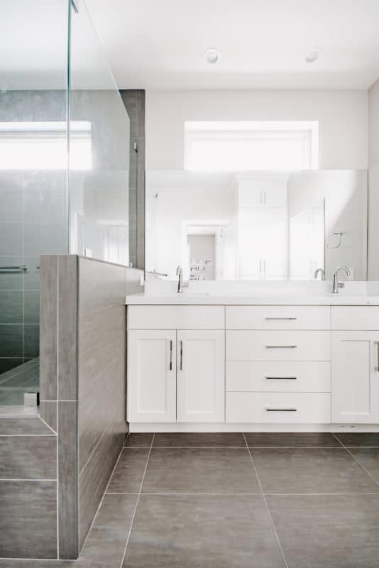 Grey tile around newly remodeled bathroom on floor and pony wall of shower, glass enclosed white cabinets and large mirror over vanity