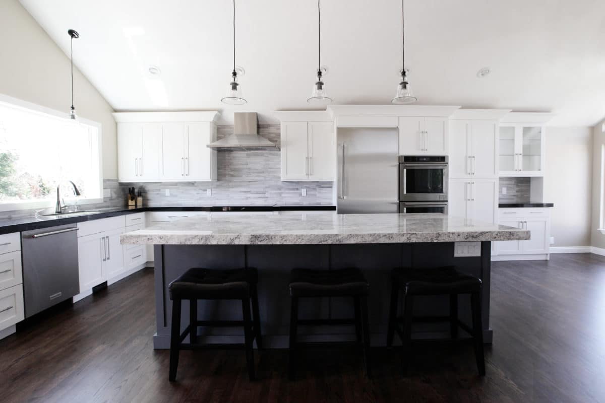 L shaped kitchen with white cabinets and dark island with pendants above. stainless steel appliances and double wall ovens