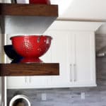 shelves over sink in kitchen holding a red colandar and bird, kitchen faucet below, shaker style white cabinets and dark countertops, stacked tile backsplash