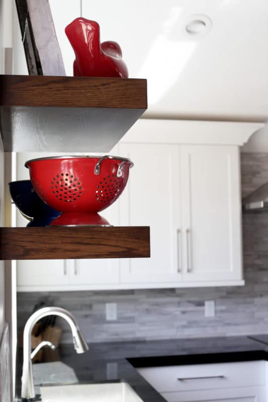 shelves over sink in kitchen holding a red colandar and bird, kitchen faucet below, shaker style white cabinets and dark countertops, stacked tile backsplash
