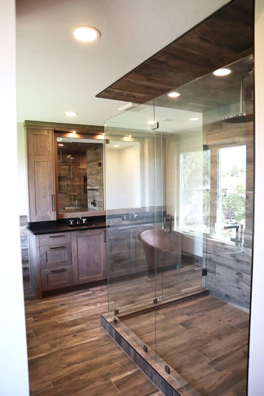 Custom home bathroom design with natural wood flooring and paneling, matching wood-finished shower, copper bathtub, dark dual sink vanity, and recessed lighting throughout space