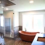 Luxurious resort feel bathroom with natural wood flooring and paneling, matching wood-finished shower, copper bathtub, dark colored countertop, dual vanity, and large window giving the space lots of natural lighting