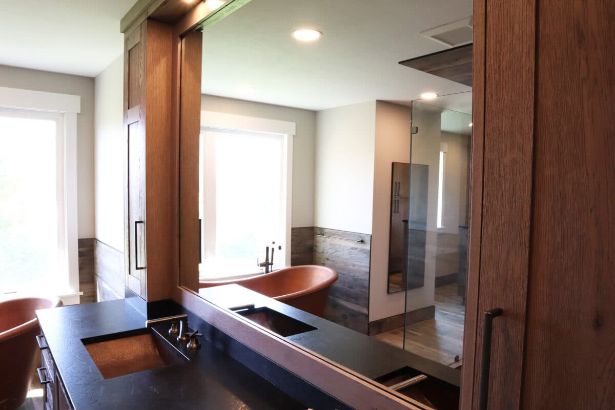 Large custom bathroom mirror with custom above and below cabinetry framing mirror, dark countertops, dual sink vanity, and copper colored bathtub.
