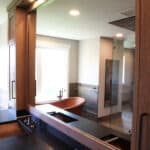 Large custom bathroom mirror with custom above and below cabinetry framing mirror, dark countertops, dual sink vanity, and copper colored bathtub.