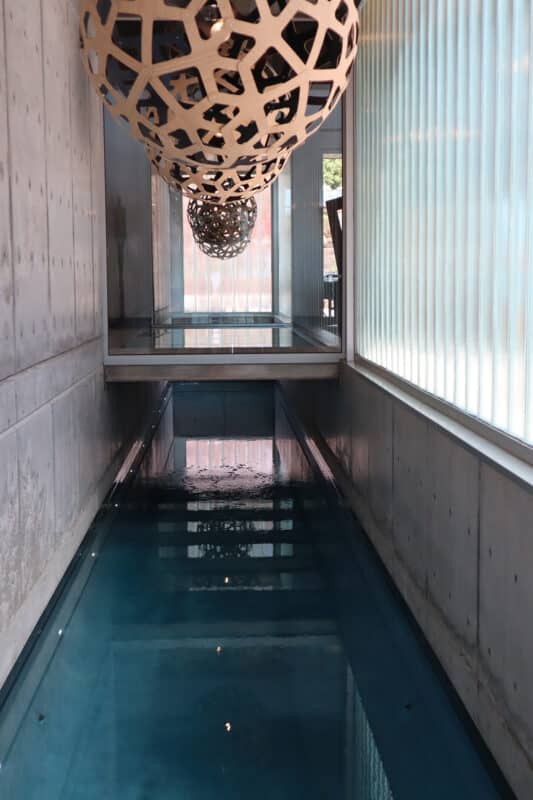 Indoor lap pool with a glass wall on the right, and a concrete wall on the left. Large Light fixture hung over the water.